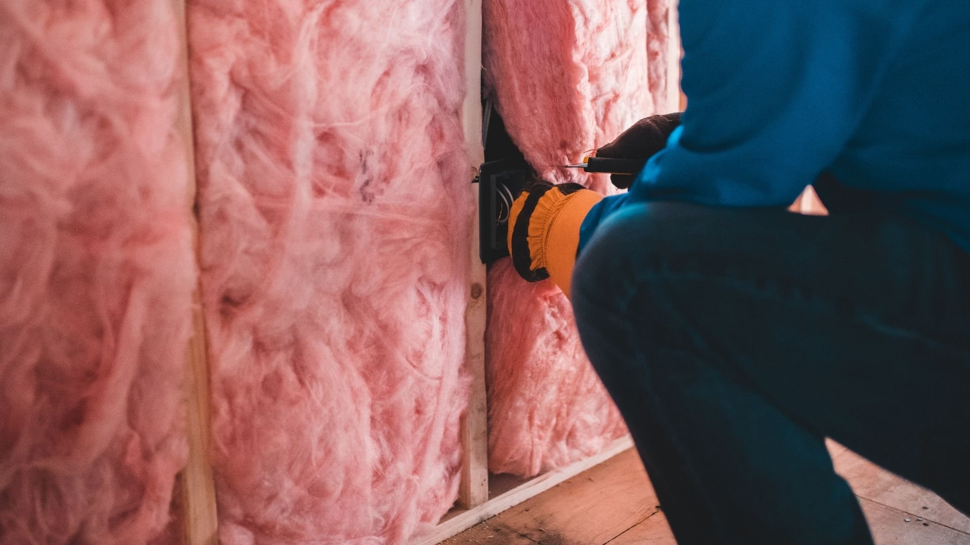 Worker in blue clothing installing pink fiberglass insulation batts in wall cavity.