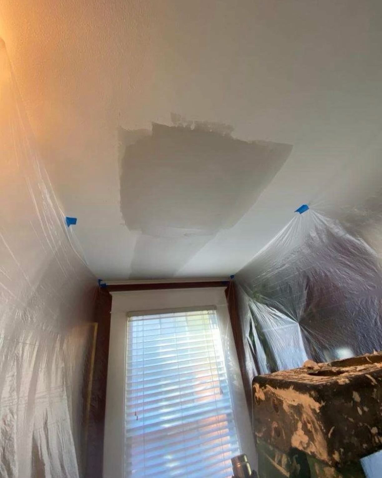 Indoor room under renovation with plastic sheeting, drywall repairs, and window visible below.